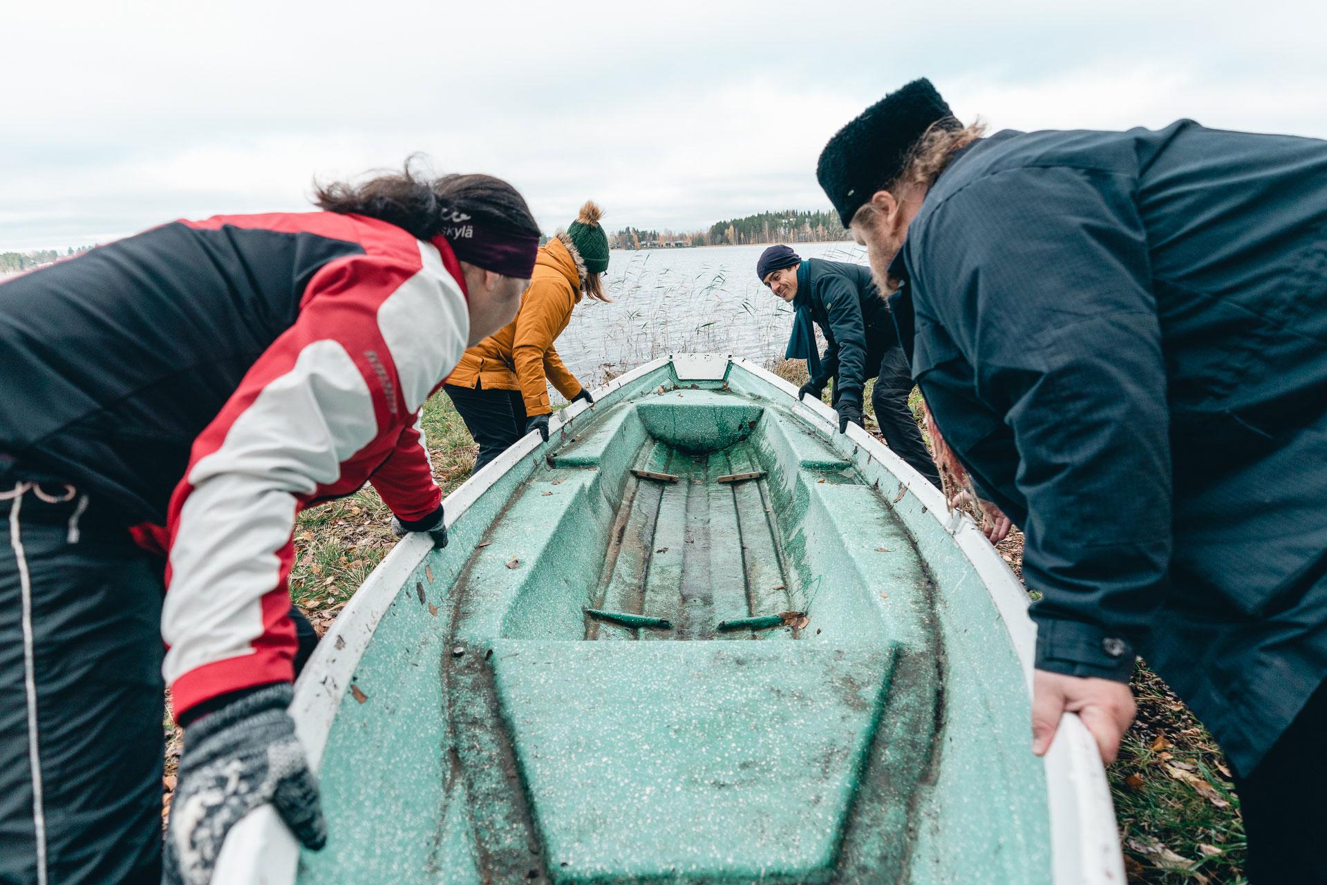 Neljä ihmistä työntää venettä vesille syksyisessä maisemassa.